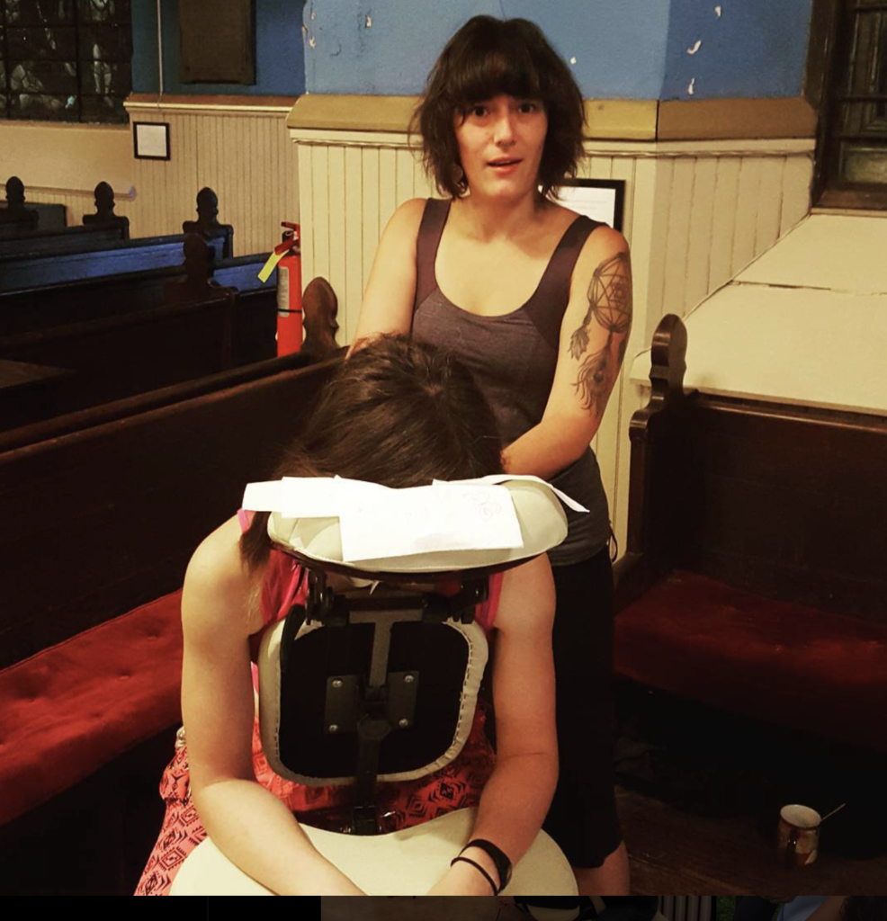 A massage therapist in a casual tank top with a visible arm tattoo standing behind a seated client in a massage chair, set in a room with church pews and stained glass windows in the background.